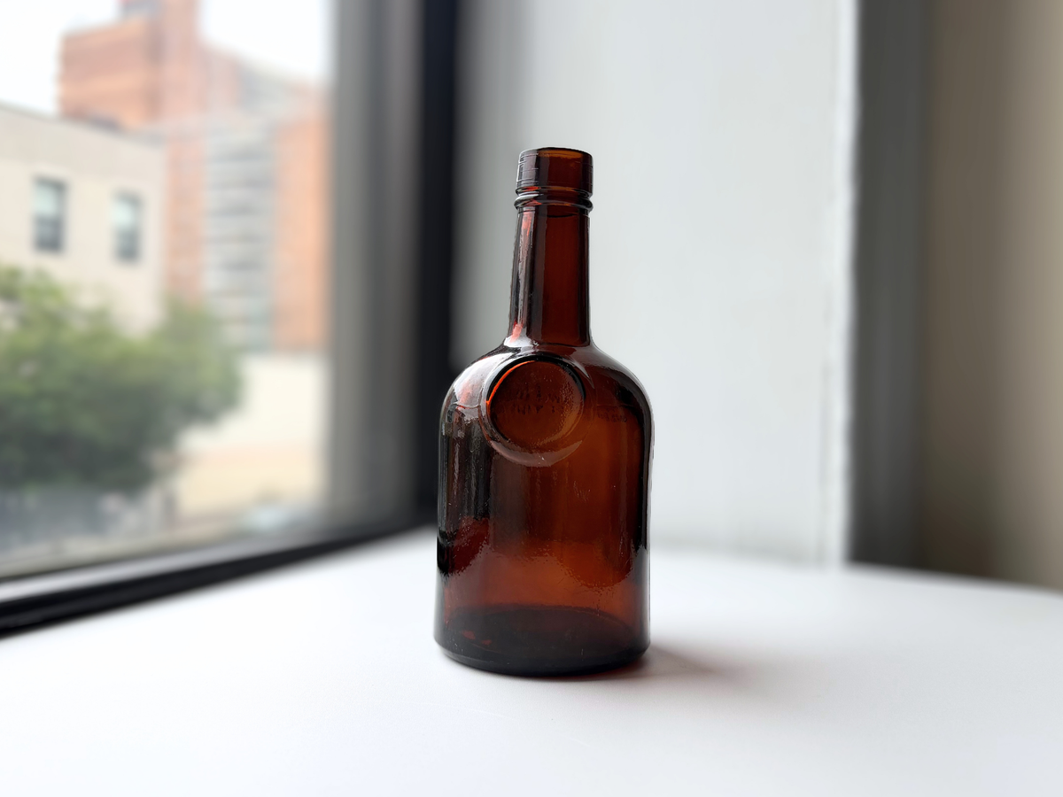 1953 dark amber glass bottle with embossed circular emblem, displayed on a windowsill.