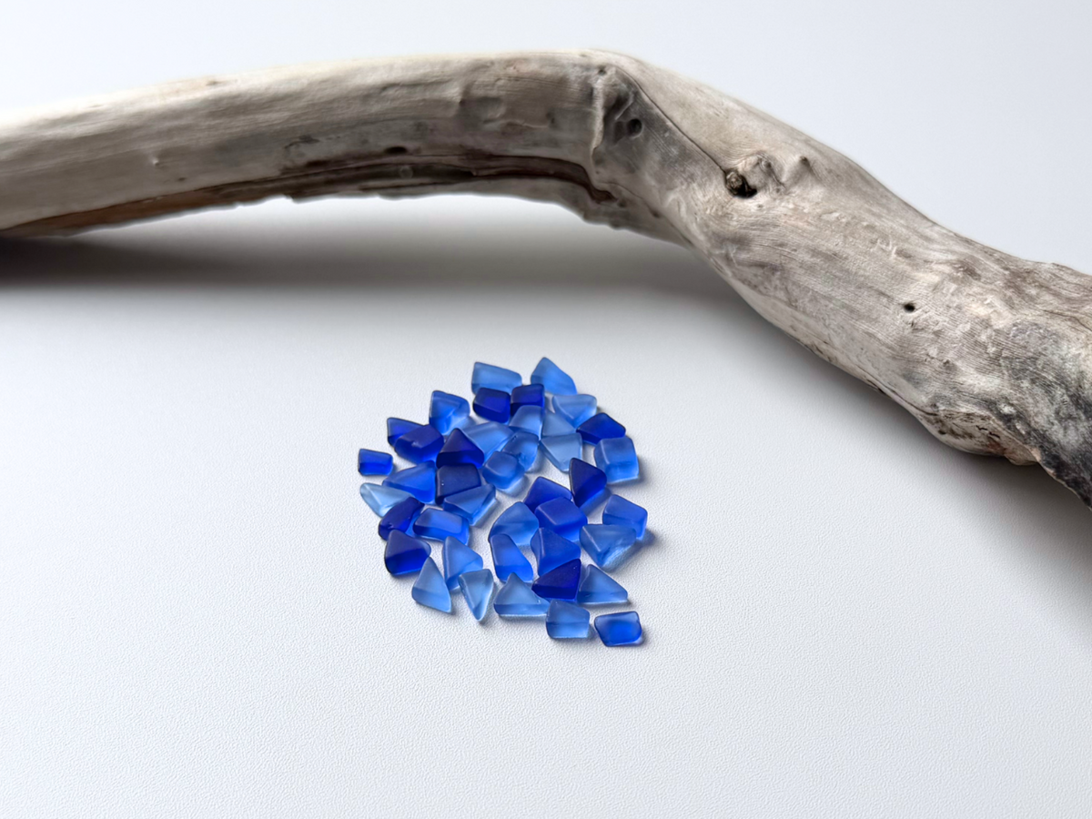 Small pile of deep blue micro tumbled sea glass resting beside a piece of driftwood on a white background, showing the tiny under-10mm size of this filler glass.