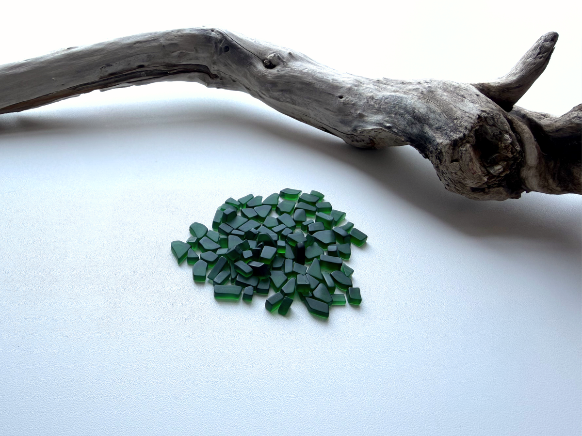Small pile of dark forest green micro tumbled sea glass resting beside a piece of driftwood on a white background, showing the tiny under-10mm size of this filler glass.