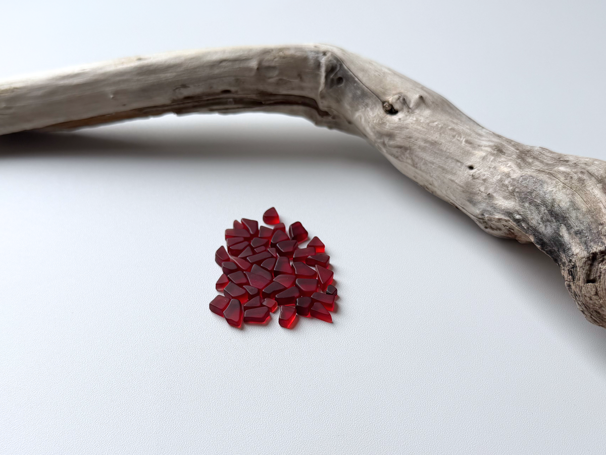 Small pile of deep cranberry red micro tumbled sea glass resting beside a piece of driftwood on a white background, showing the tiny under-10mm size of this filler glass.