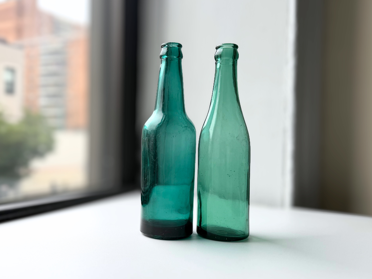 Pair of teal glass beer bottles, a 1950s Danish Kastrup bottle and an antique Hamilton Glass Co. bottle, displayed together on a windowsill.