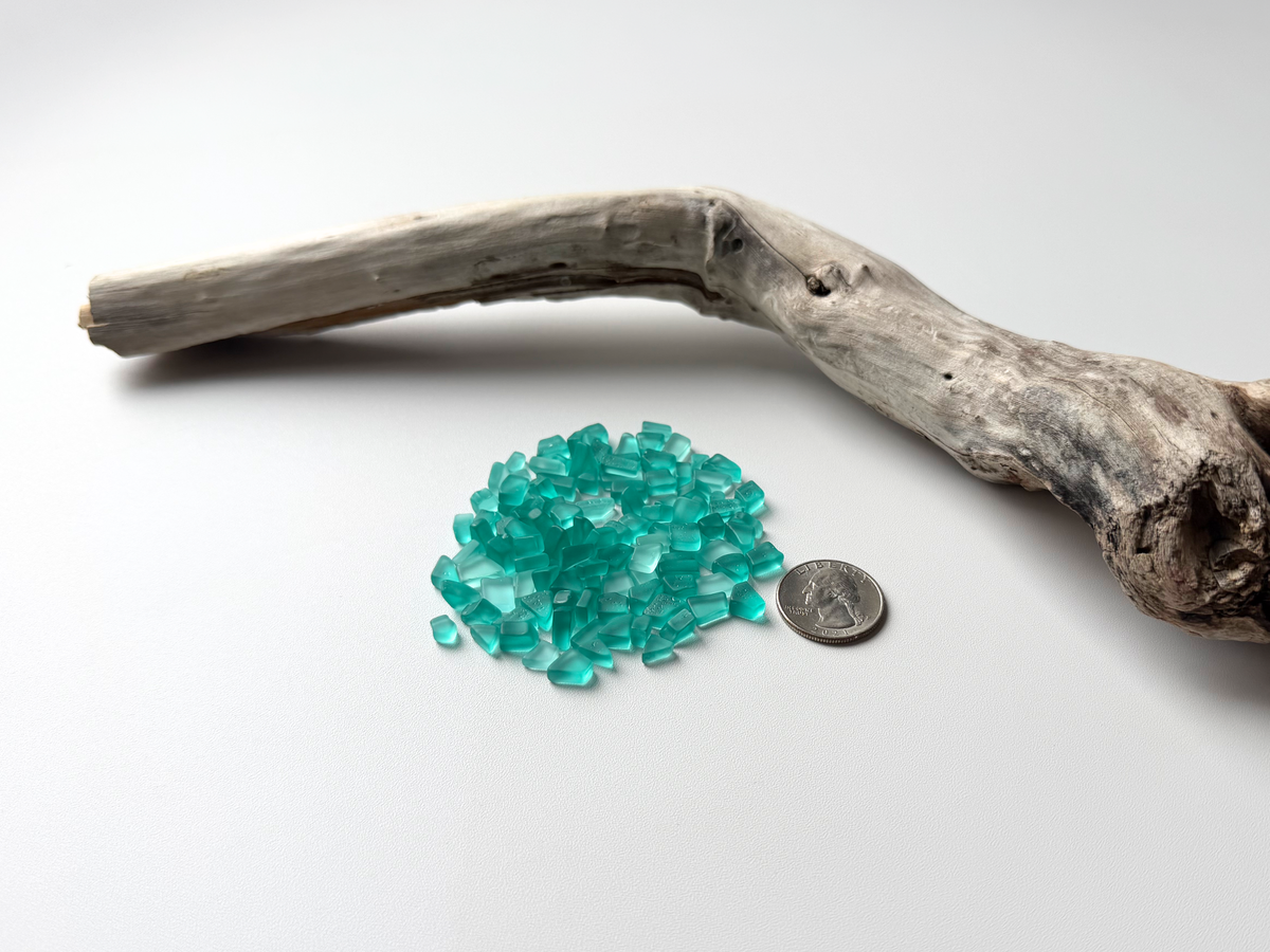 Aqua micro sea glass pile next to a coin for size comparison, on driftwood background.