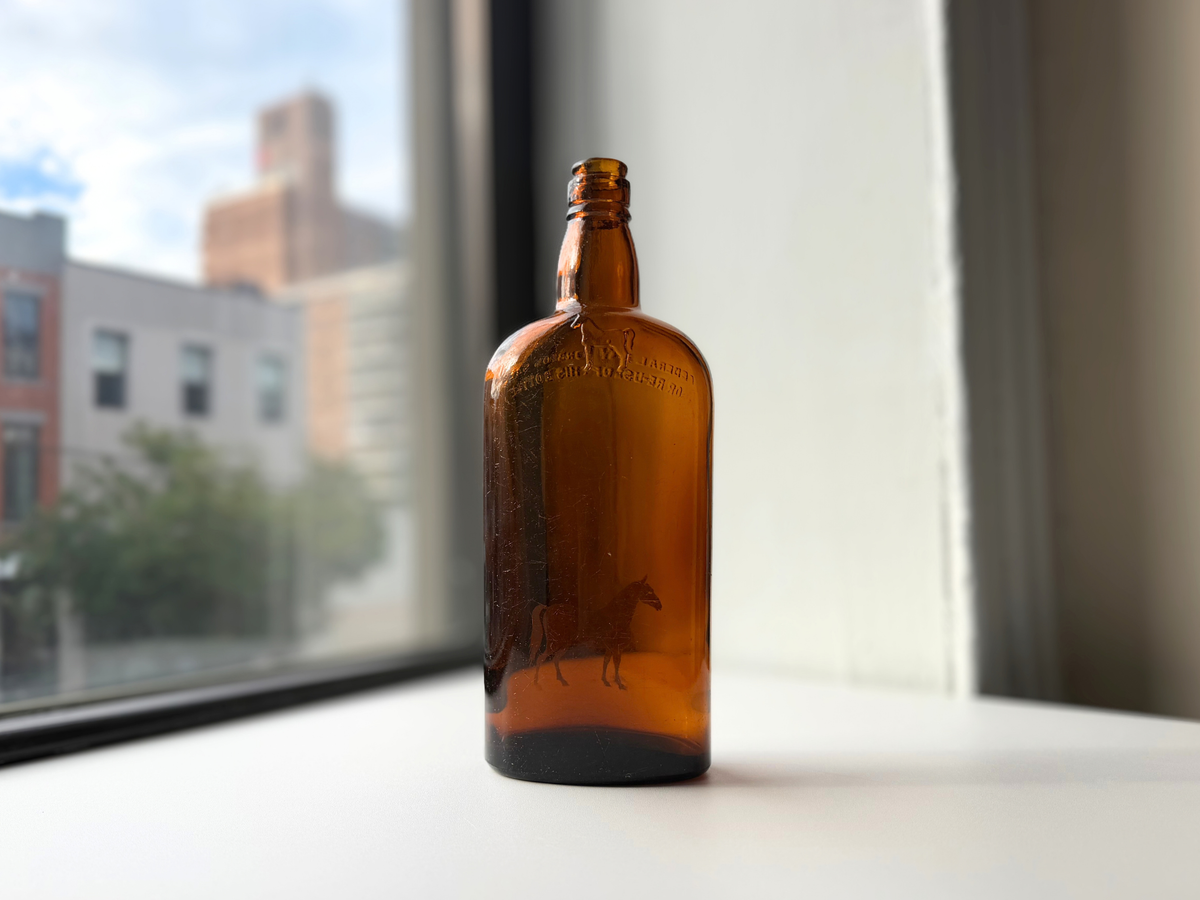 Back of the vintage amber White Horse bottle on a windowsill, with the horse and the embossed shoulder lettering showing through the glass in reverse.