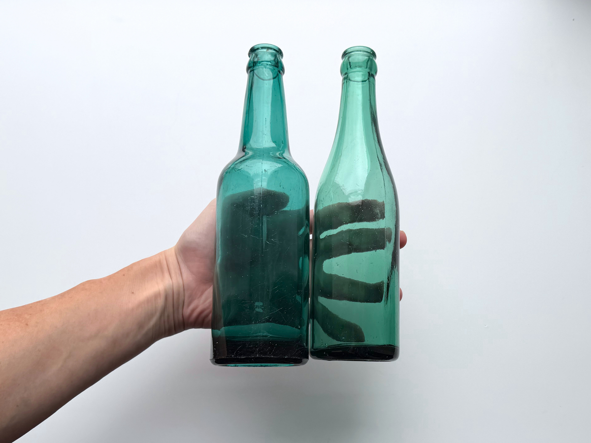 Hand holding two teal glass beer bottles together to show their matching size and color.