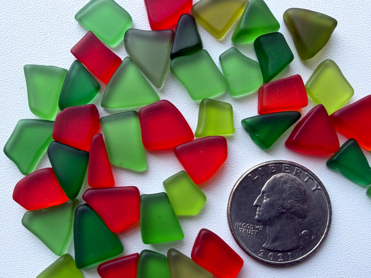 Close-up of holly-red and holiday-green tumbled sea glass pieces scattered next to a quarter for size reference, illustrating the tiny .25 to .50 inch pieces included in this Christmas ornament mix.