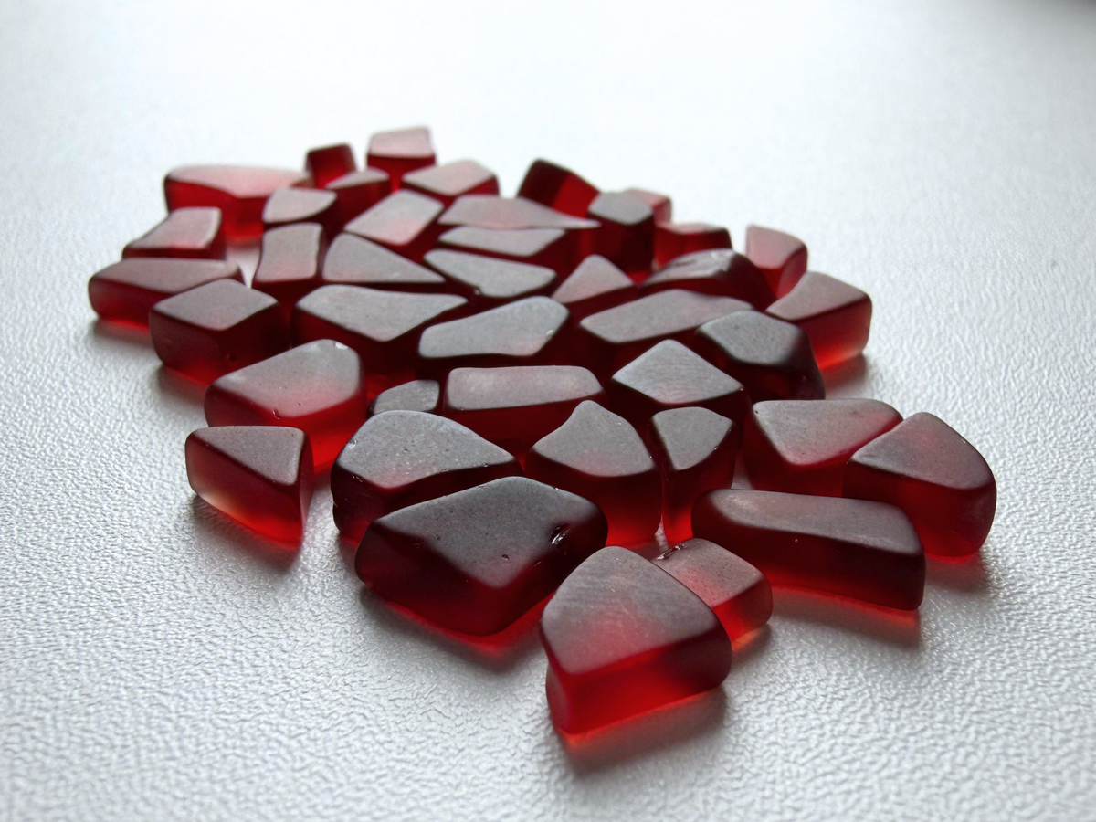 Abundant pile of cranberry red micro sea glass filler pieces spread across a white background, showing the deep red color and small tumbled shapes.
