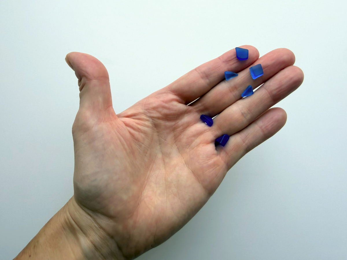A hand holding a small handful of deep blue micro tumbled sea glass pieces, giving a human-scale sense of just how tiny this filler glass is.