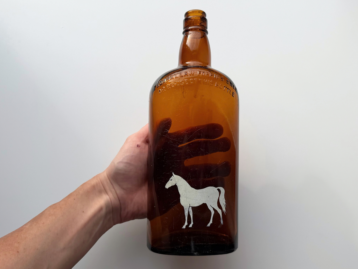 Hand holding the vintage amber White Horse bottle to show its size, with the painted white horse on the front and embossed lettering on the shoulder.