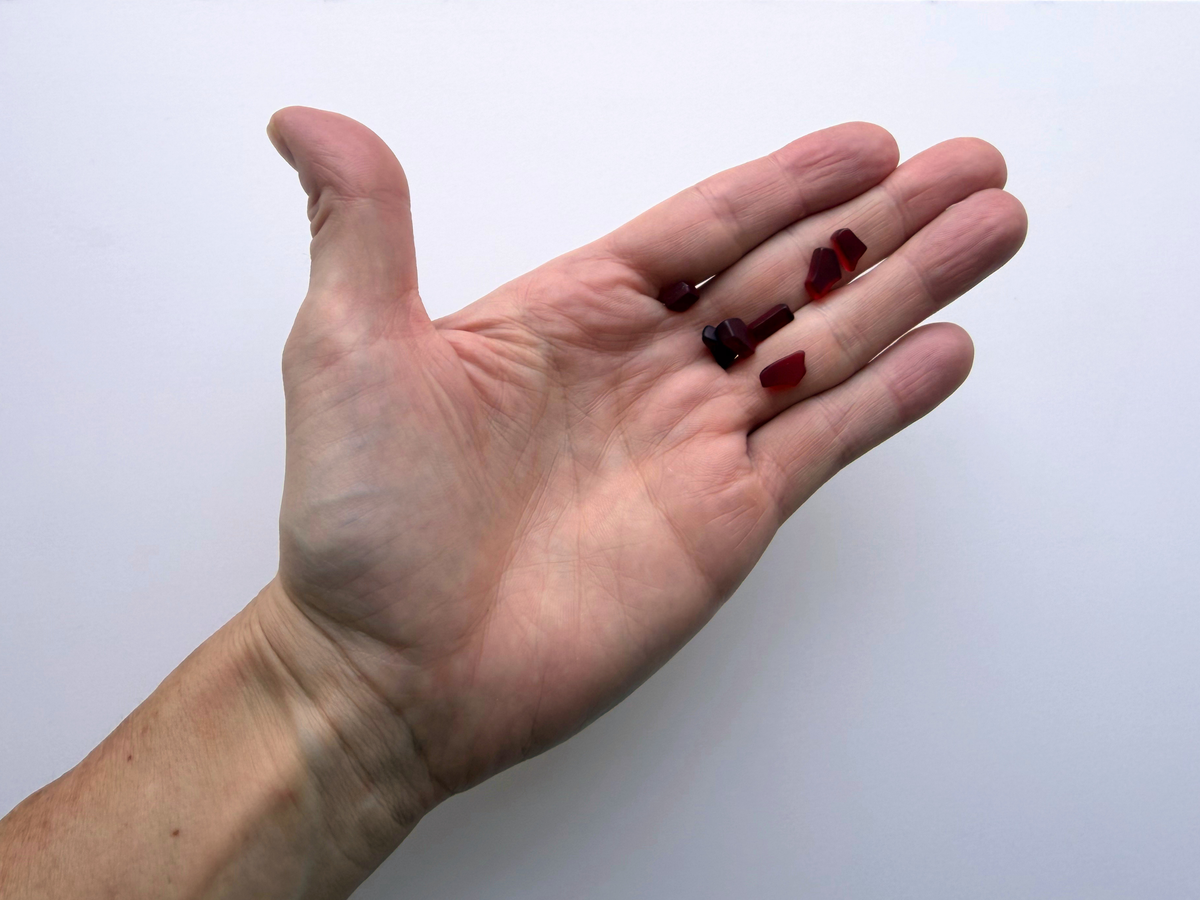 A hand holding a small handful of cranberry red micro tumbled sea glass pieces, giving a human-scale sense of just how tiny this filler glass is.