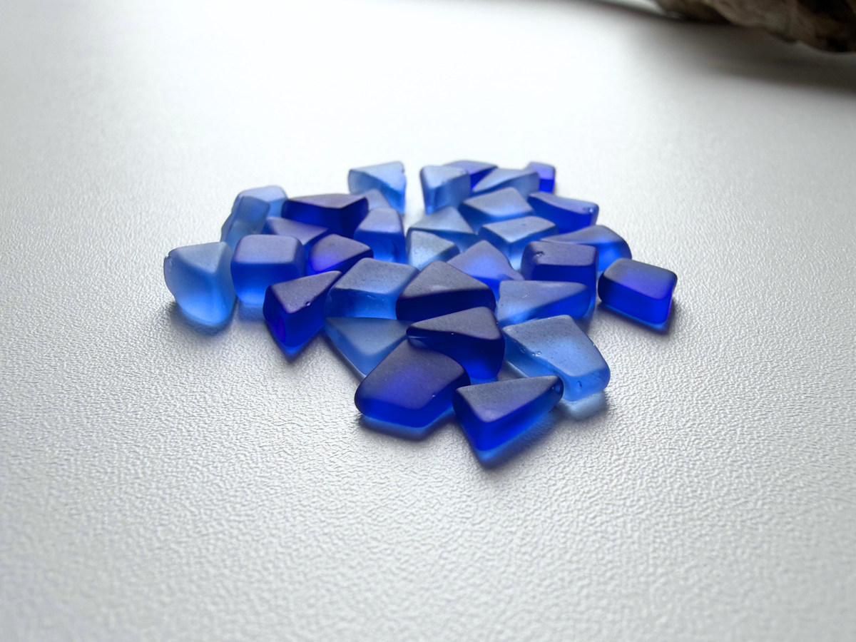 Abundant pile of deep blue micro sea glass filler pieces spread across a white background, showing the periwinkle-to-cobalt color range and small tumbled shapes.