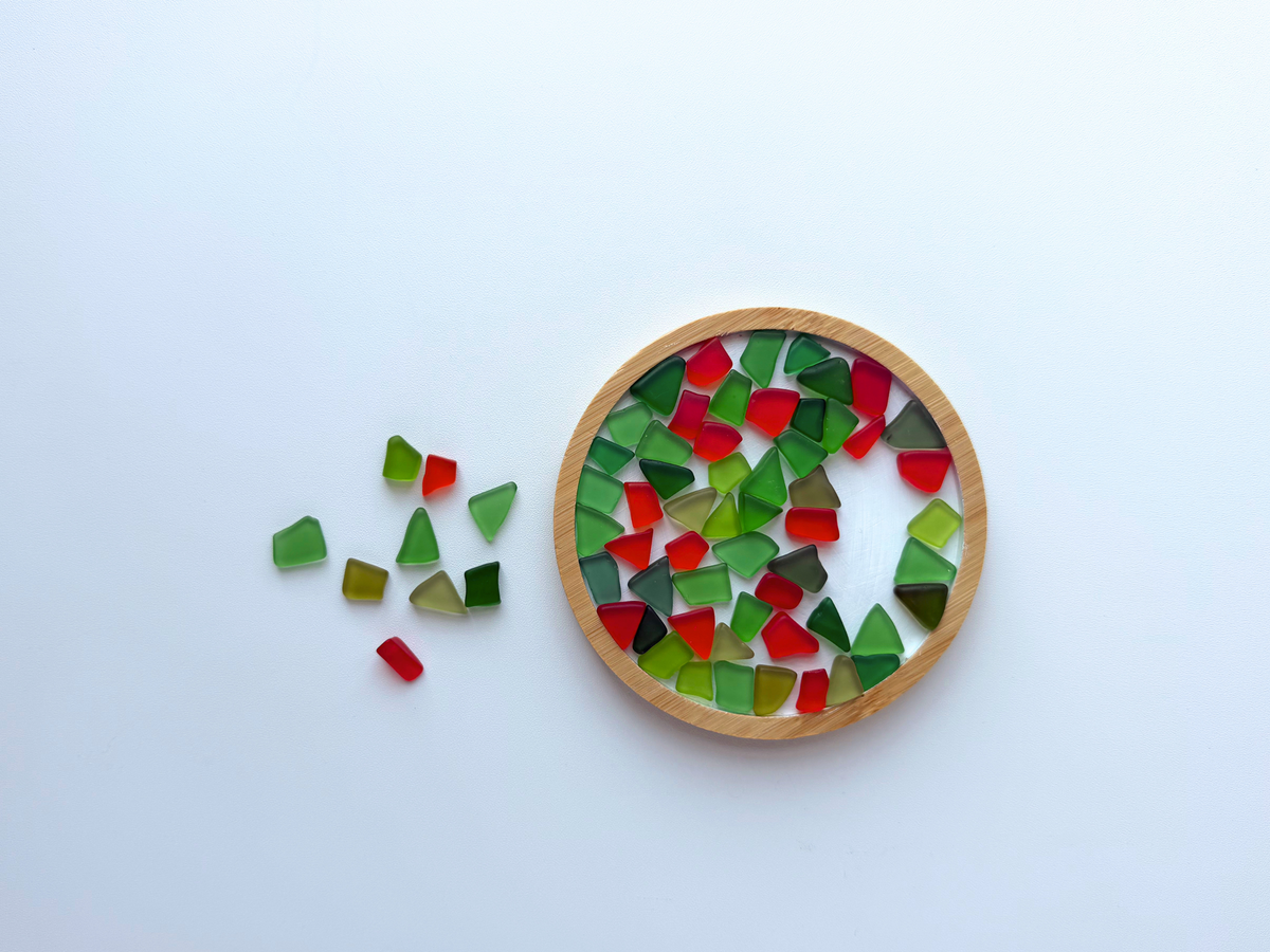 Holly-red and green tumbled sea glass pieces spilling out beside a small round wooden dish filled with the same glass, styled to show the mix used as a decorative holiday accent or catch-all bowl filler.