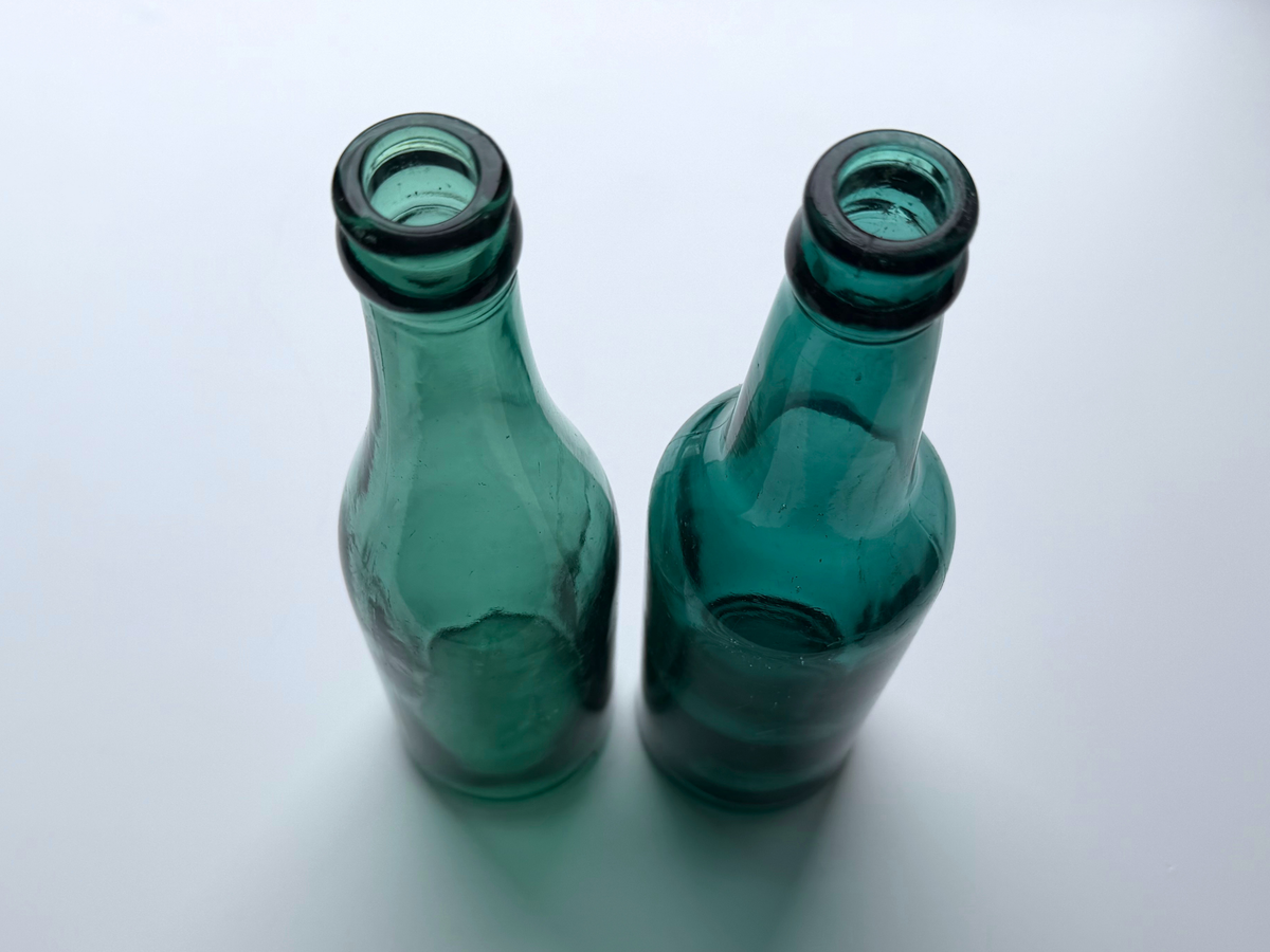 Close view of the open mouths and necks of two teal glass beer bottles against a gradient background.