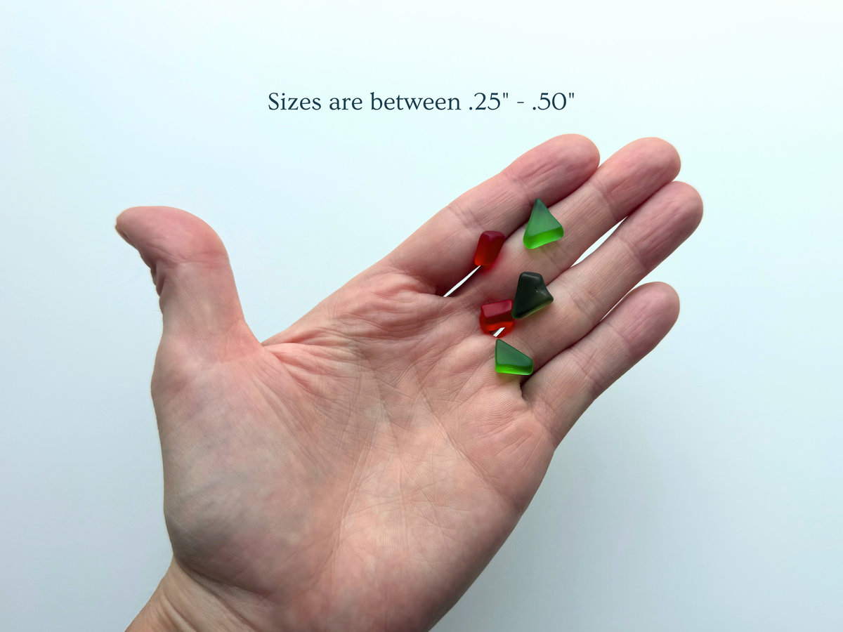 A hand holding a few holly-red and green tumbled sea glass pieces with text overlay noting sizes range between .25 and .50 inches, giving buyers a true sense of scale before purchase.