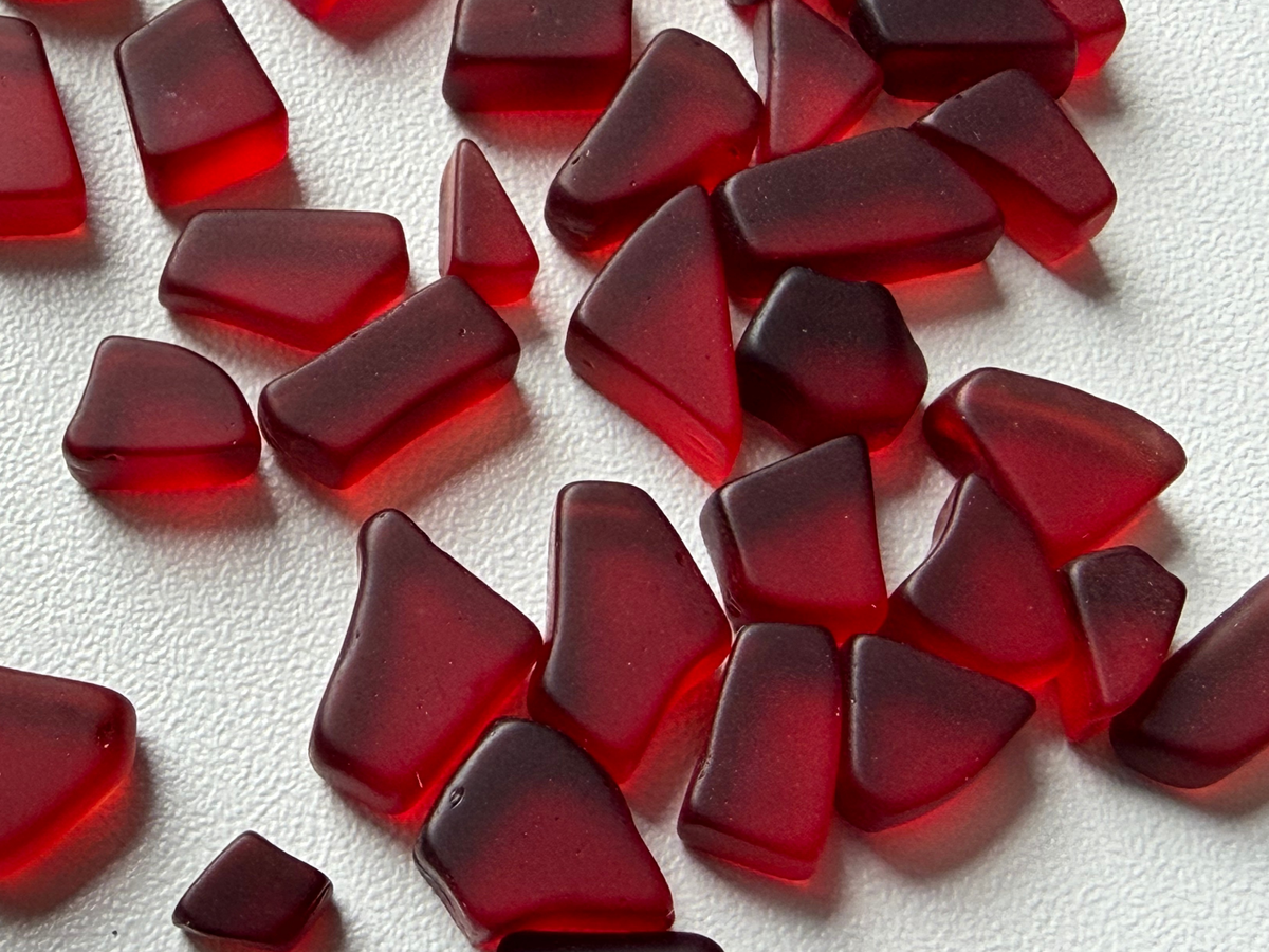 Second angle of a loose pile of cranberry red micro tumbled sea glass, showing the range of shapes and consistent deep red color in this filler mix.