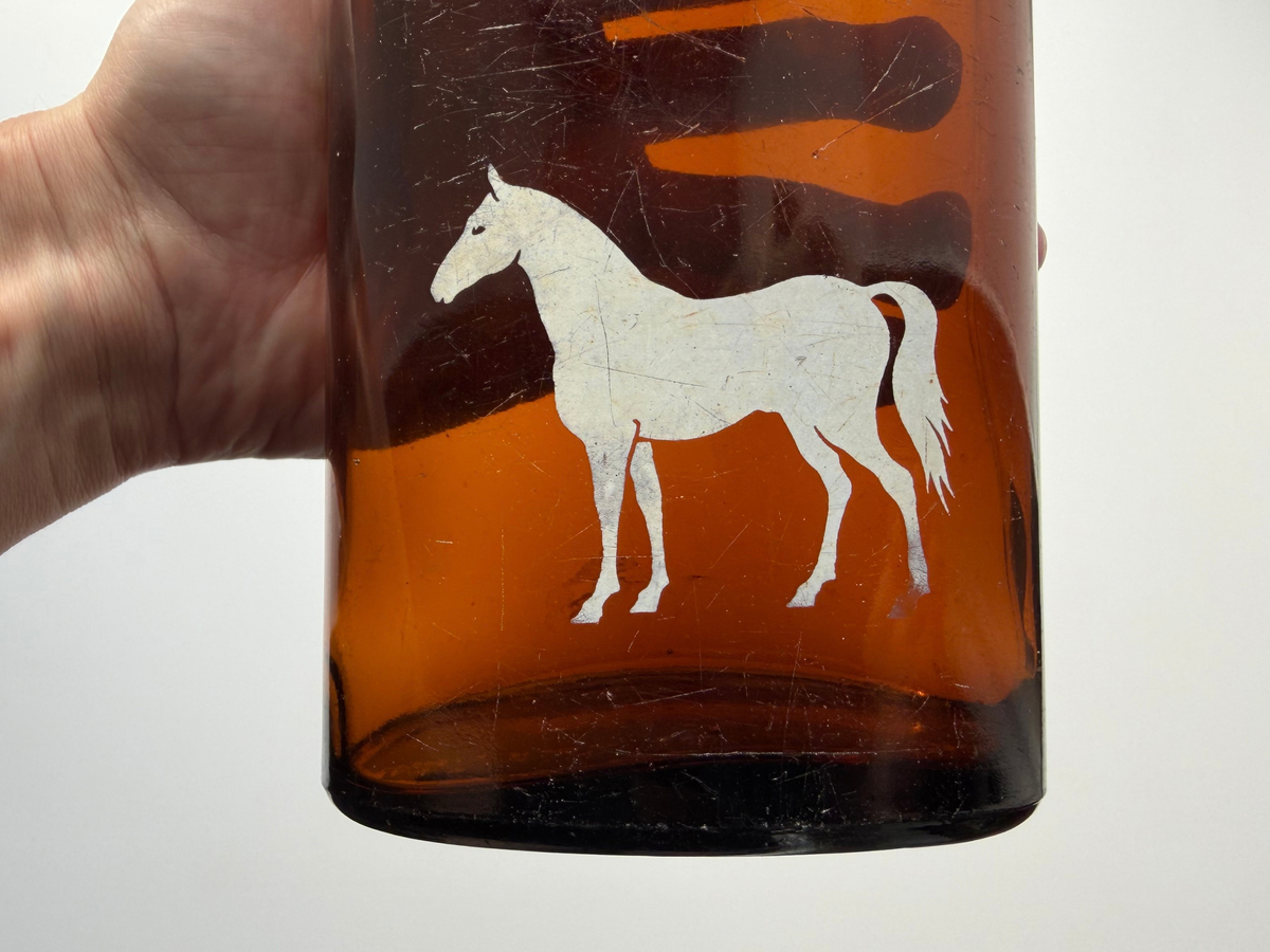Close-up of the painted white horse on the front of a vintage amber glass bottle, showing light surface scratches in the glass and the paint.