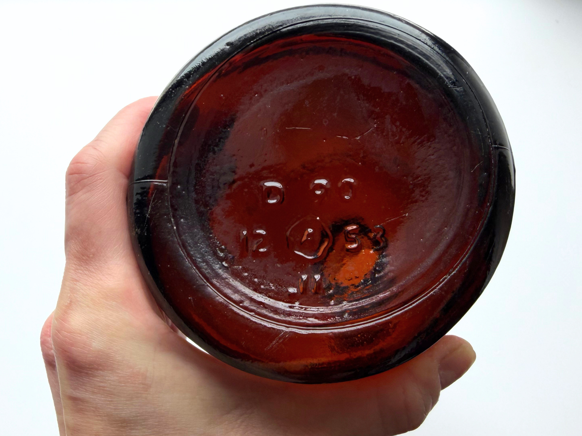 Dark amber glass bottle base held in hand showing embossed circular emblem, dating it to 1953.
