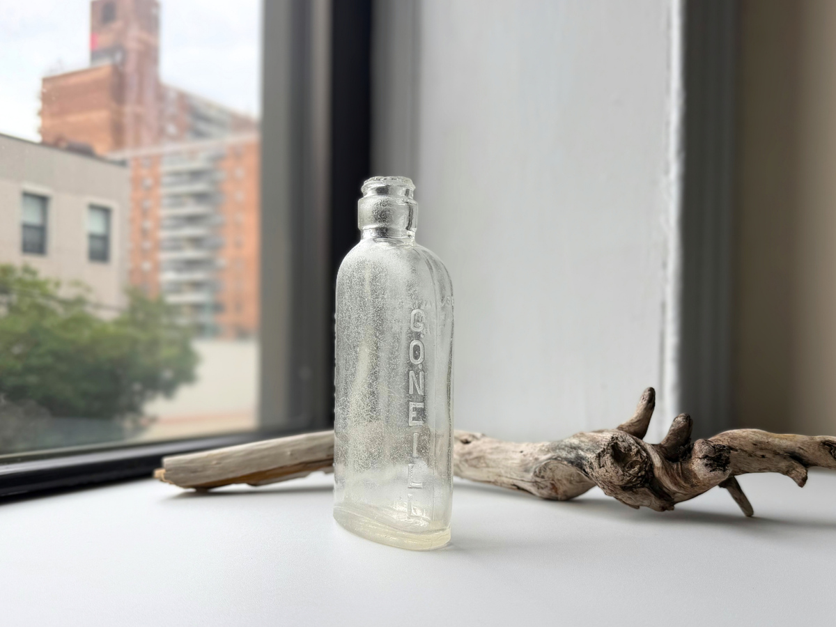 Side profile of a clear sea-worn G. O'Neill flask on a windowsill with G.ONEILL embossed vertically and a rounded, frosted mouth.