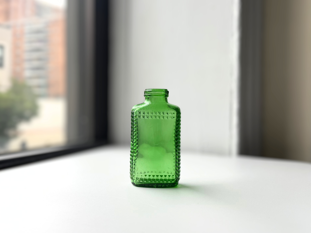 Vintage kelly green Airwick hobnail glass bottle on a windowsill, angled view