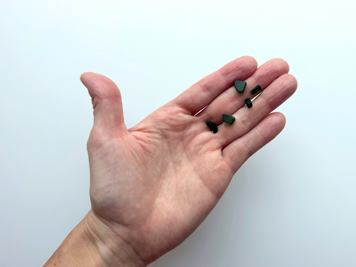 A hand holding a small handful of dark green micro tumbled sea glass pieces, giving a human-scale sense of just how tiny this filler glass is.