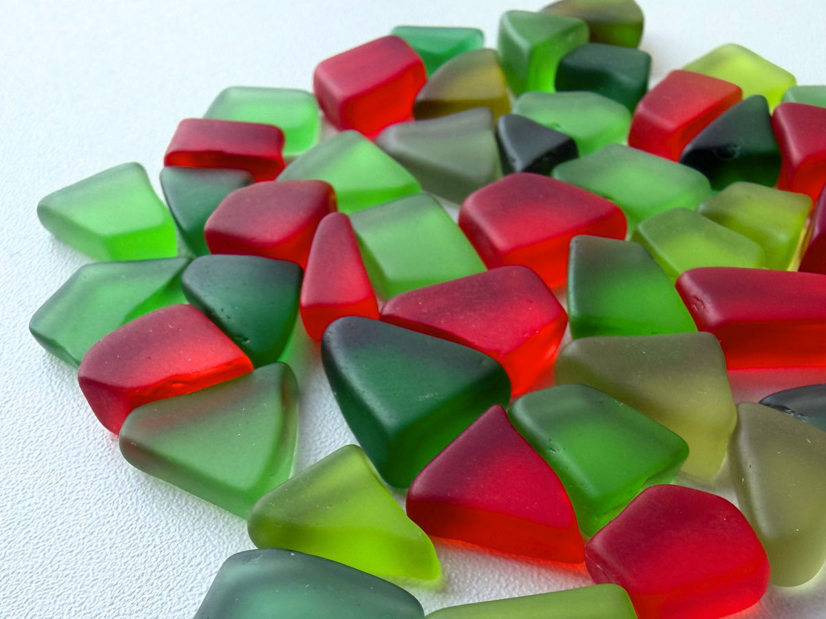 Full top-down view of an abundant pile of holly-red and holiday-green tumbled sea glass pieces on a light textured background, showing the range of shades included in each Christmas ornament mix set.