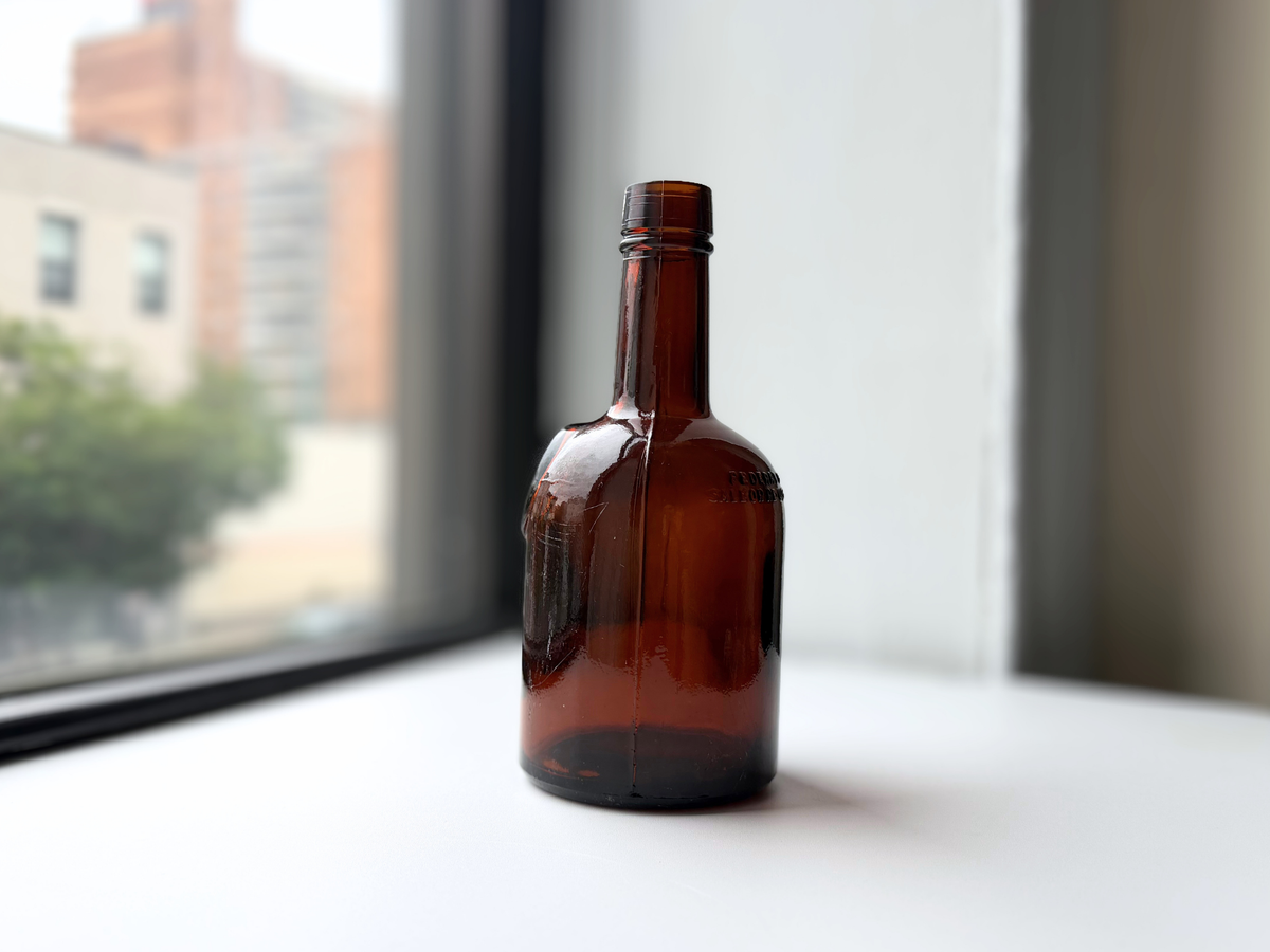 1953 dark amber glass bottle displayed on a windowsill with Brooklyn brownstones in the background.