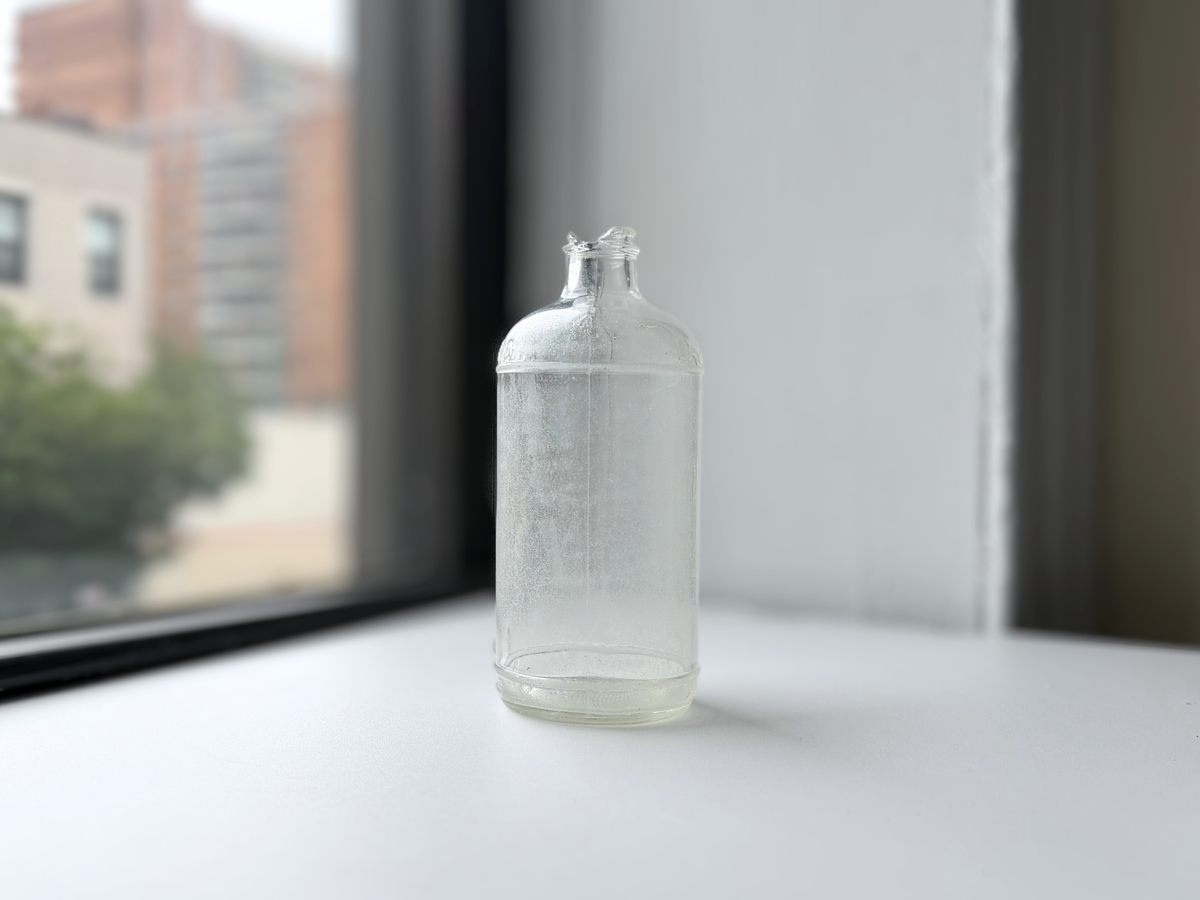 Sea-worn frosted glass bottle on a sunlit windowsill with a Brooklyn building in the background.