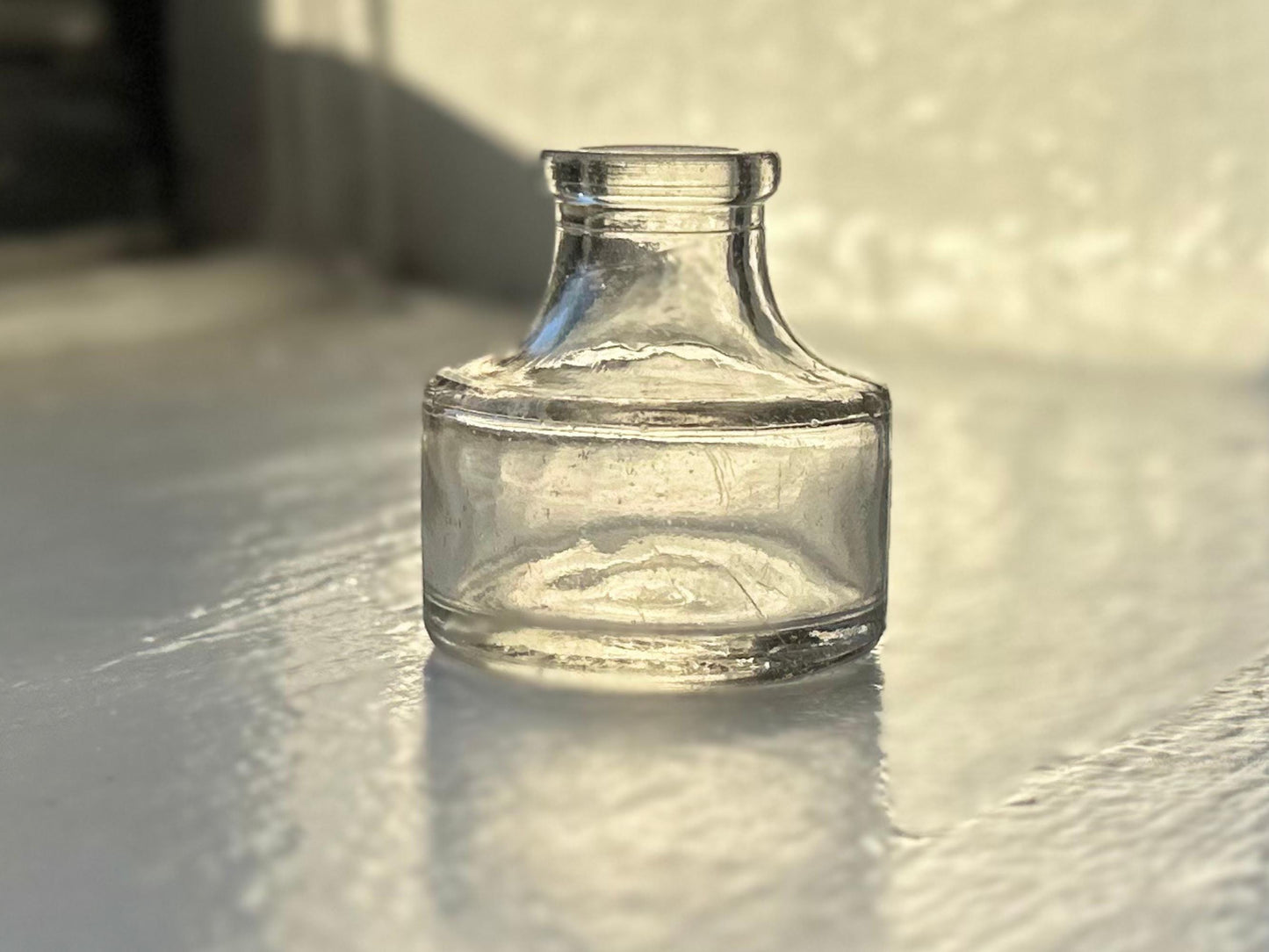 A small, antique Higgins Ink Jar from 1947. The jar is clear glass with a cylindrical shape and a short neck. It sits on a white surface with a slight reflection.