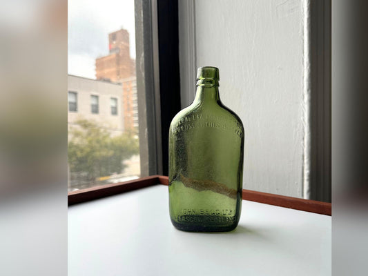 Front view of a green glass John Begg Ltd. whiskey bottle embossed with “GLASGOW SCOTLAND” and a federal Prohibition warning, recovered from a NYC landfill. Vintage patina and character-rich wear make it a perfect accent for fall bar decor, antique displays, or rustic autumn vignettes.