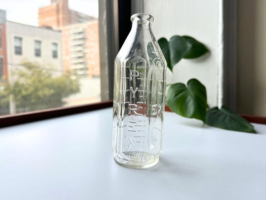 Front view of vintage 1950s Pyrex baby bottle showing embossed vertical PYREX text on clear glass, 6.4 inches tall. Mid-century nursery decor bottle perfect for cottagecore or minimalist styling. Rescued vintage glassware from Brooklyn Sea Glass for sustainable home decor. Use as bud vase for single stem flowers or display with other clear glass bottles. American-made post-war collectible ideal for baby shower gifts or gender-neutral nursery displays. Excellent condition with no chips.