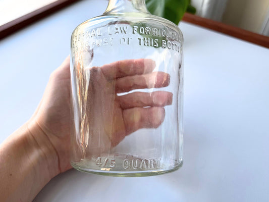 Vintage 4/5 quart clear glass liquor bottle 9.5 inches tall by 3.75 inches diameter, rescued from NYC beaches. Features embossed "FEDERAL LAW FORBIDS SALE OR RE-USE OF THIS BOTTLE" and "4/5 QUART" markings from the regulated spirits era 1935-1960s. Authentic mid-century American liquor bottle perfect for bar cart decor, vintage spirits collections, or tall single stem vases. Beach-rescued bottle with age-appropriate clarity.