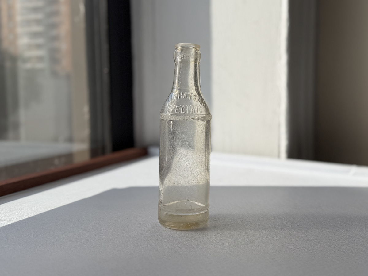 Manhattan Special espresso soda bottle full front view by window, clear embossed glass vintage NYC