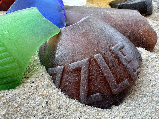 Frosted brown sea glass piece with embossed lettering resting in sand beside green and blue pieces