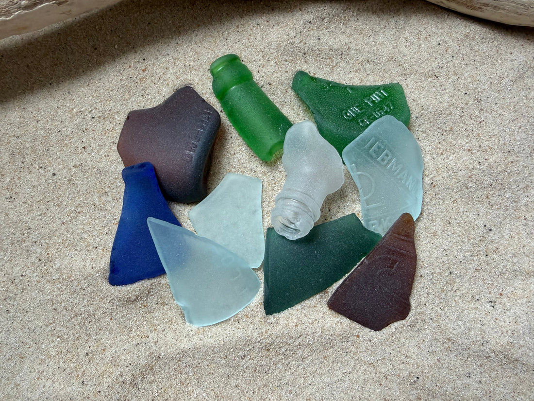 Ten frosted sea glass pieces in cobalt, green, teal, brown and clear scattered on sand, some with embossed lettering