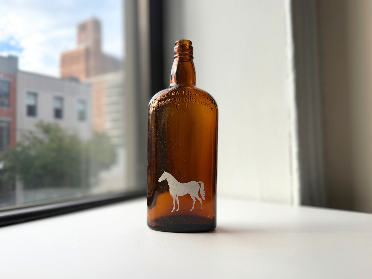 Vintage amber glass White Horse bottle with a painted white horse on the front, standing on a sunny windowsill.