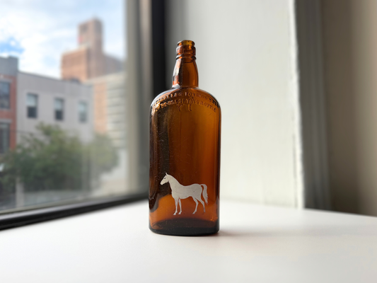Vintage amber glass White Horse bottle with a painted white horse on the front, standing on a sunny windowsill.