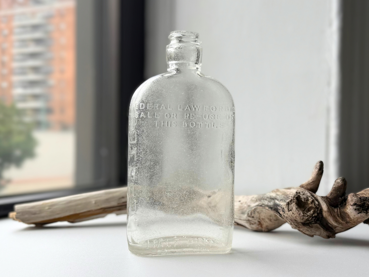 Clear sea-worn G. O'Neill flask with Federal Law embossing on a white windowsill beside driftwood, city buildings in the window behind.