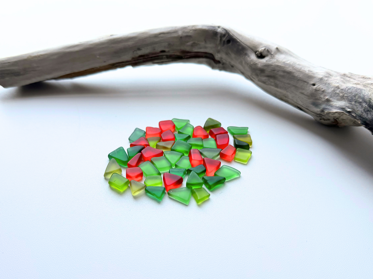 Small pile of red and green holly-colored tumbled sea glass resting beside a piece of natural driftwood on a white background, showing the mix's petite size for Christmas ornaments and holiday crafts.