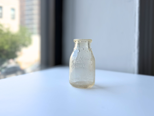 Sea-worn clear glass Elmhurst Cream Co dairy bottle on a sunlit windowsill, full front view showing genuine sea glass patina