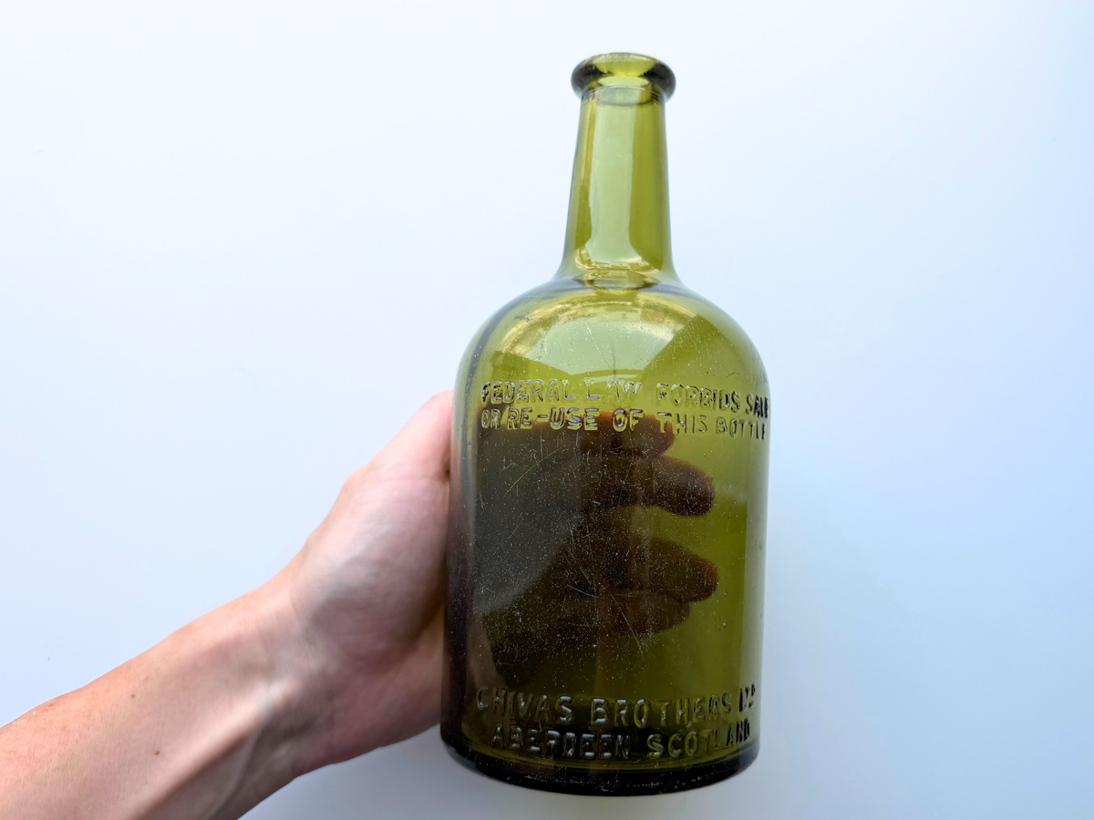 Hand holding a vintage light olive green Chivas Brothers glass bottle to show its scale