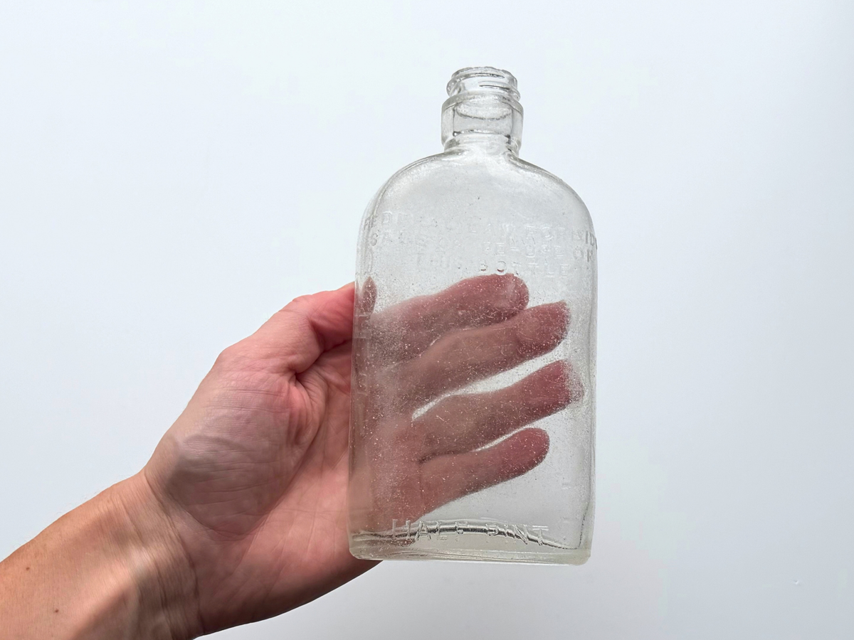 Hand holding a clear sea-worn G. O'Neill half pint flask against a white background, showing the frosted surface and embossed lettering.