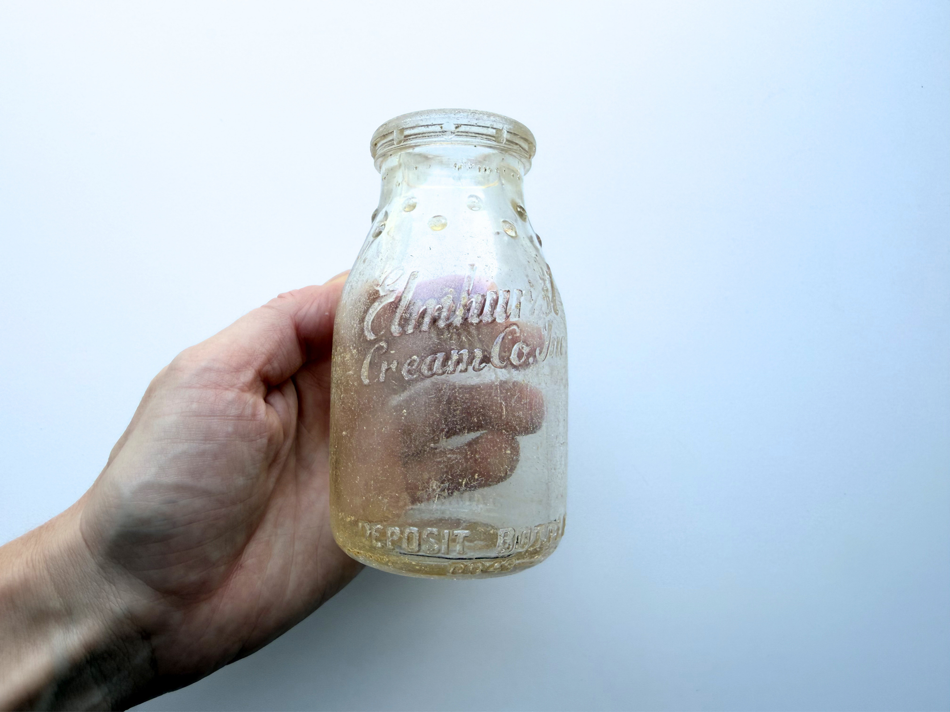 Hand holding sea-worn Elmhurst Cream Co glass dairy bottle, showing embossed lettering up close
