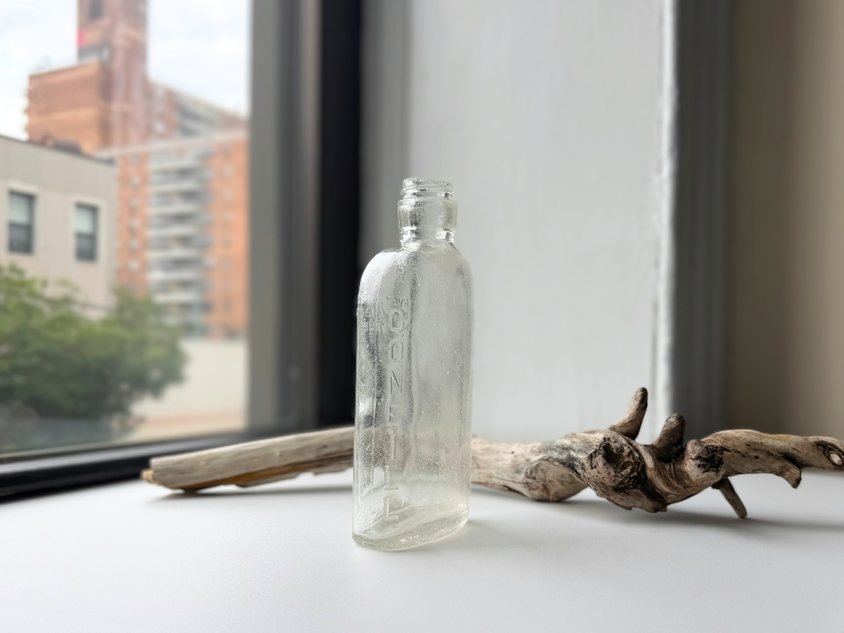 Angled view of a clear sea-worn G. O'Neill flask on a windowsill, showing vertical G.ONEILL embossing on the narrow side next to driftwood.