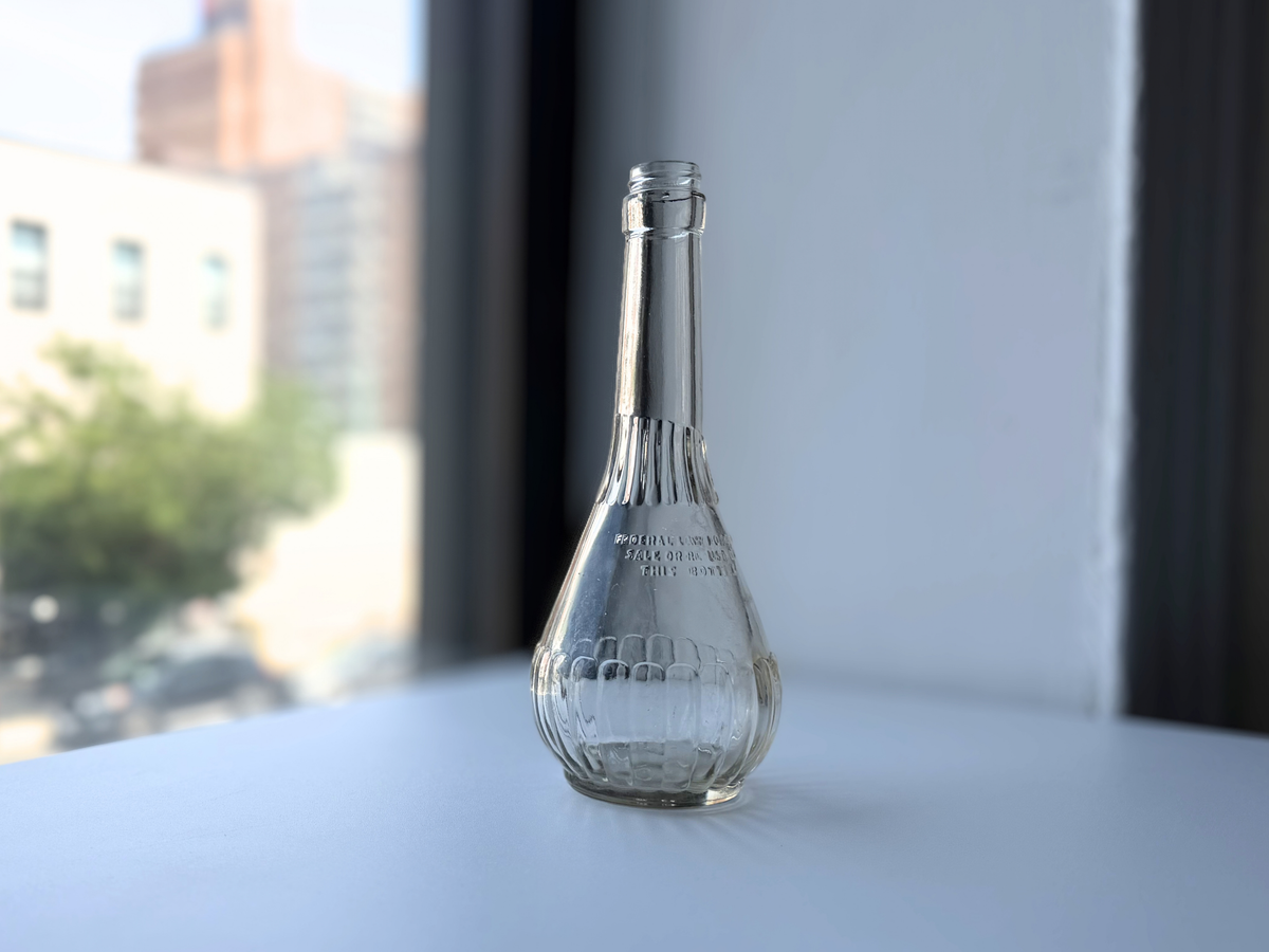 Angled view of a vintage clear glass genie bottle with a ribbed base and tall neck
