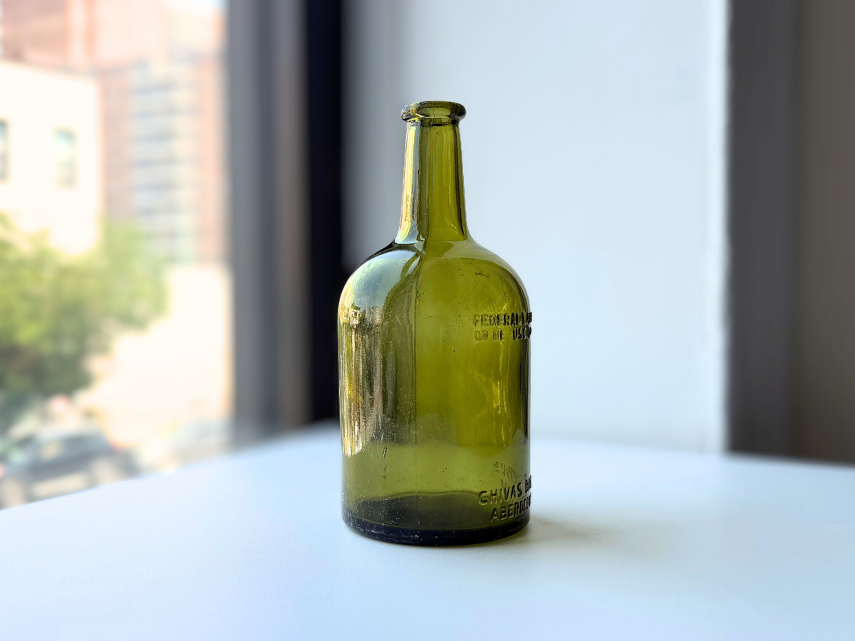 Angled side view of a vintage olive green glass Chivas bottle with embossed lettering