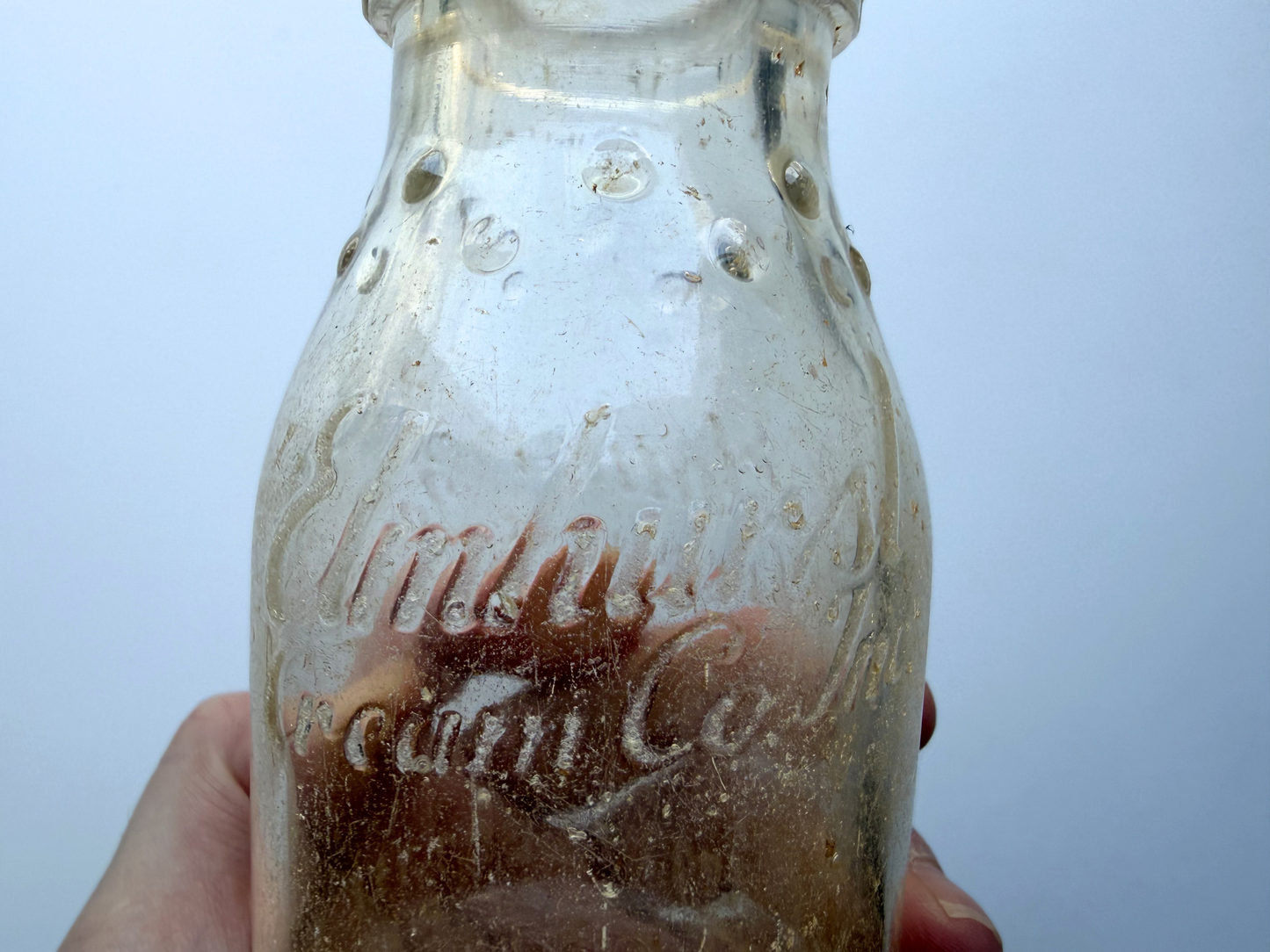 Close-up of frosted embossed lettering reading Elmhurst Cream Co on a sea glass patina bottle held in hand