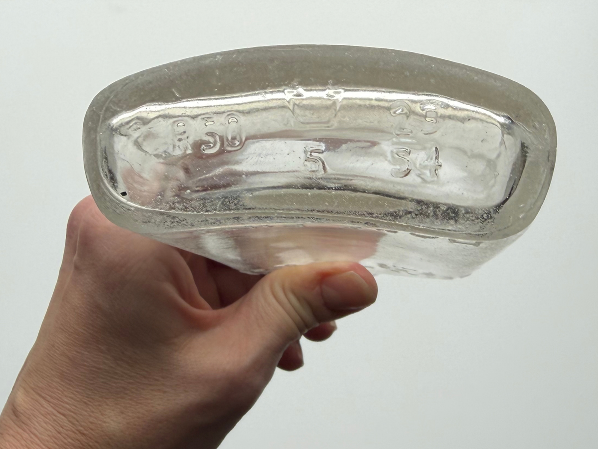 Hand holding a clear sea-worn G. O'Neill flask base-up, showing the embossed base markings including the date number 54.