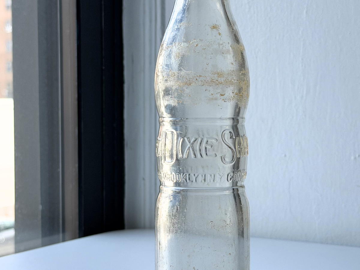 Close-up of a vintage Dixie Shake bottle on a windowsill highlighting the embossed DIXIE SHAKE lettering and surface patina.