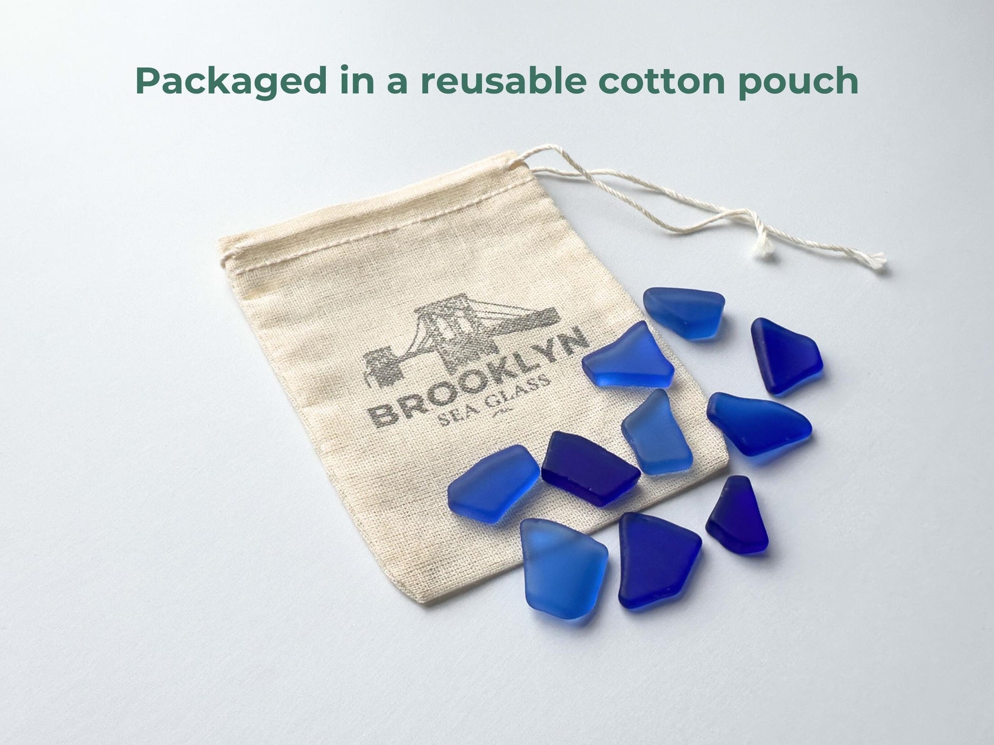 Cobalt blue tumbled sea glass spilling from a Brooklyn Sea Glass–branded reusable cotton pouch on a white background. Eco-friendly, recycled from vintage glass, and smoothed for safe crafting. Ideal for jewelry making, mosaics, resin art, beach weddi