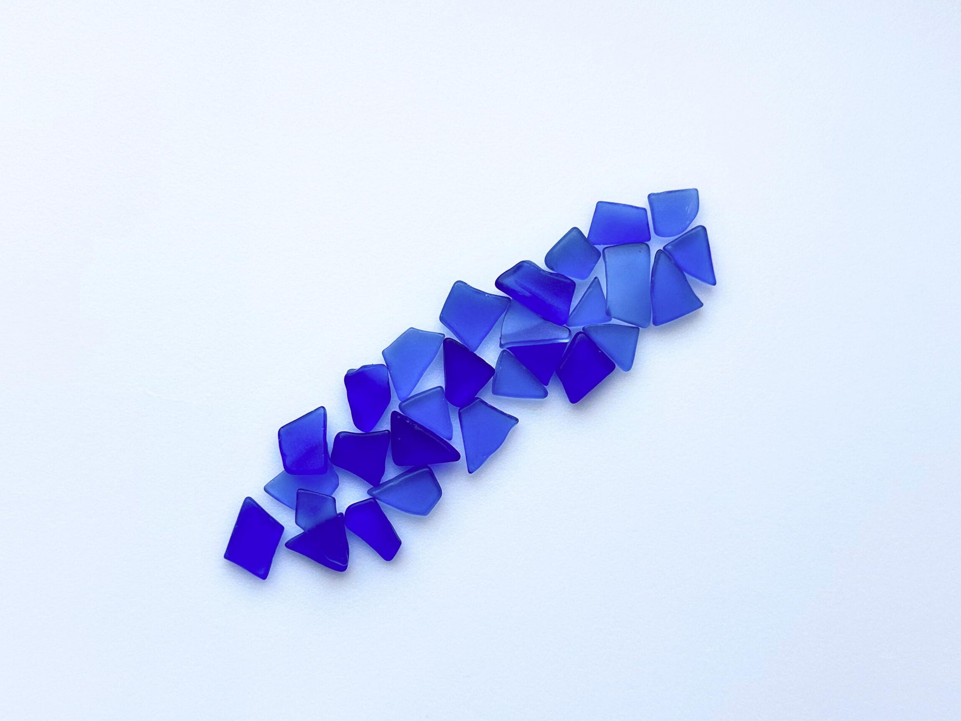 Flat lay of deep blue tumbled sea glass in a scattered line on white background – perfect for jewelry and resin art