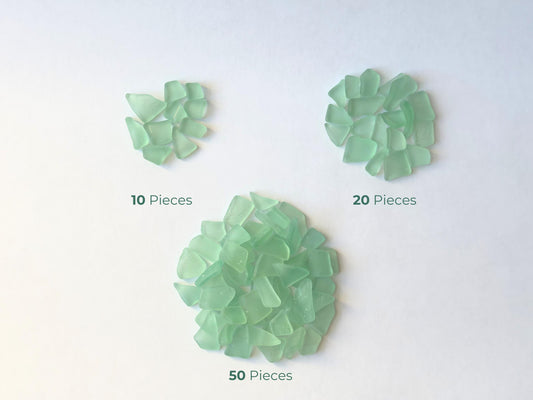 Grouped display of 10, 20, and 50 pieces of seafoam green tumbled glass, machine-finished to mimic ocean-polished sea glass. Made from vintage Coca-Cola bottles collected along NYC shores, these shards are smooth, frosted, and size-sorted—perfect for